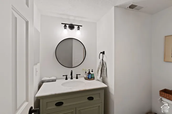 Olive-toned vanity featuring a white countertop with an integrated basin, a matte black three-handle faucet, a circular mirror with a black frame, and a three-light fixture with clear glass shades