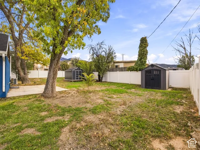 Backyard area featuring a concrete patio, white vinyl fencing, two detached storage sheds, and mature trees with a mountain backdrop