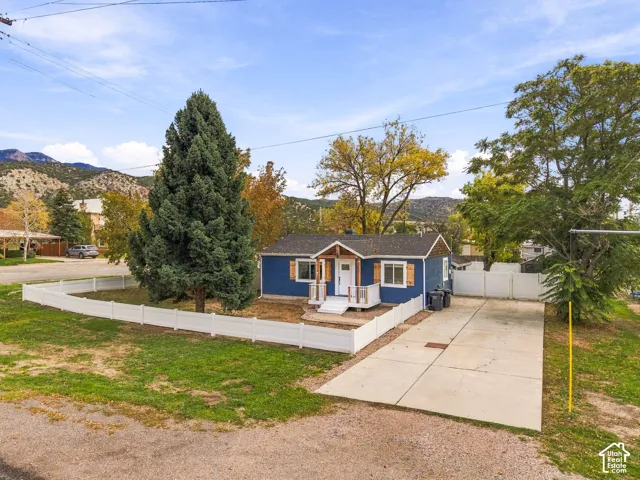 Exterior featuring blue siding, front porch with wood accents, concrete driveway, white vinyl fencing, and mature landscaping against a mountain backdrop
