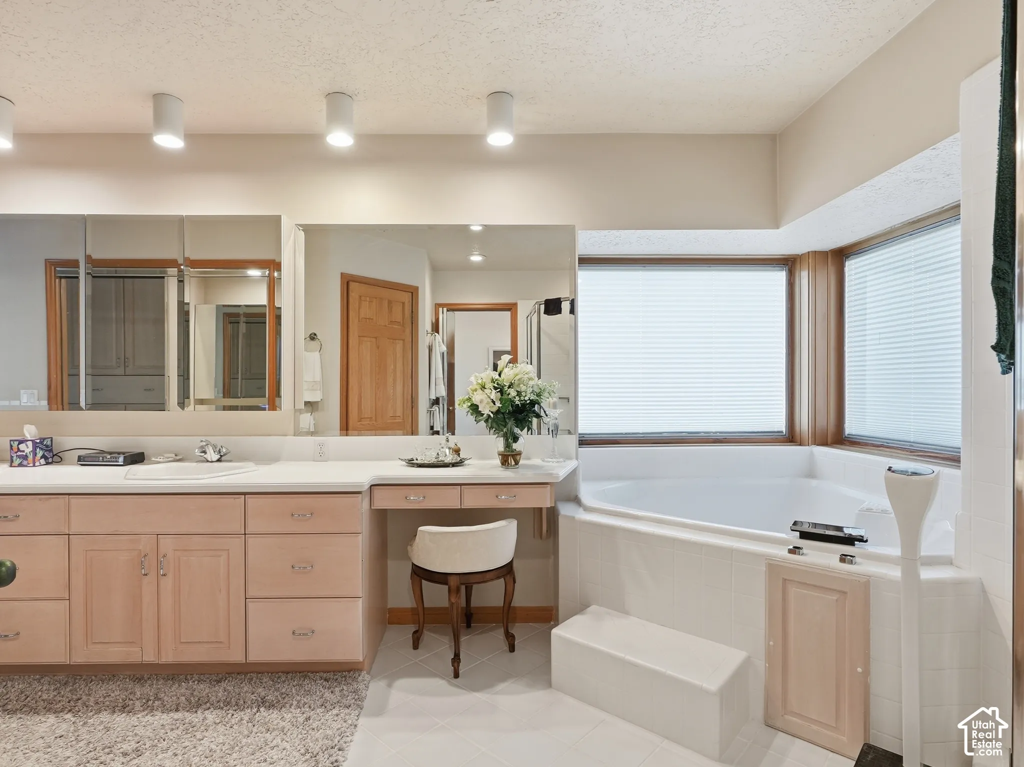 Bathroom vanity featuring light wood cabinetry, integrated dressing table, expansive wall-to-wall mirror, and tiered corner soaking tub with surround tiling
