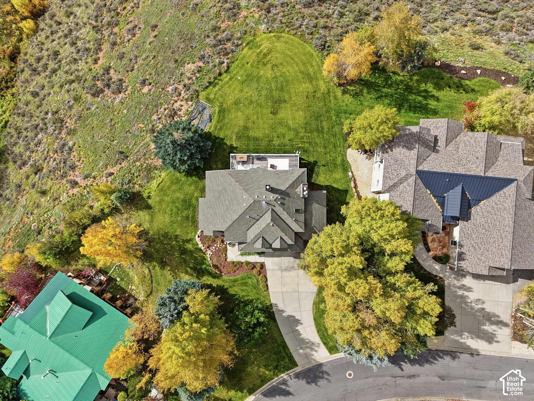 Aerial perspective of a multi-gabled residence featuring a concrete driveway, an elevated deck, extensive lawn space, and mature trees