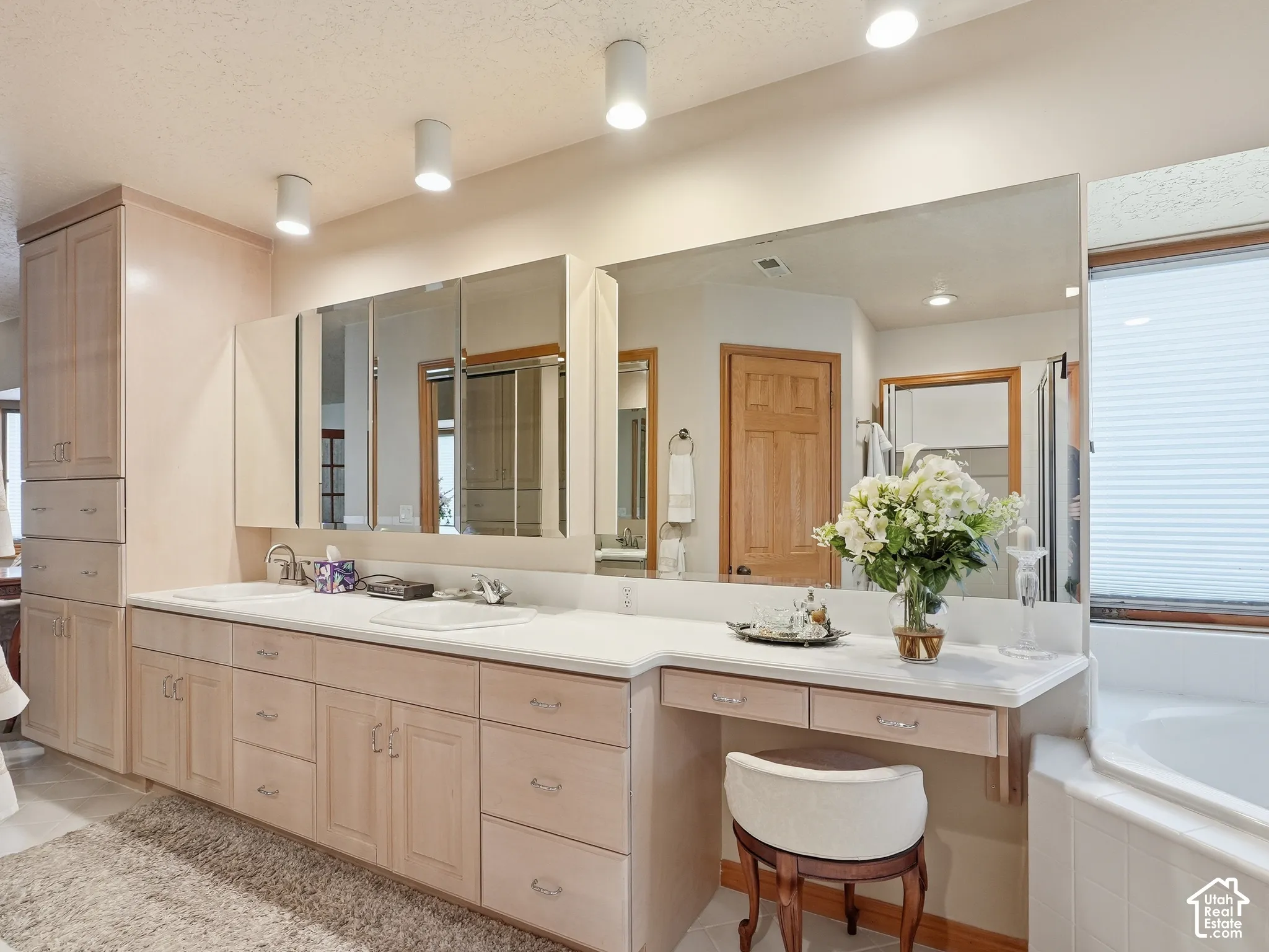 Dual-basin vanity featuring light-finish cabinetry, built-in storage tower, inset sinks, integrated vanity seating area, and wall-to-wall mirror