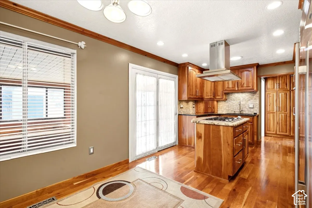 Kitchen featuring wood cabinetry, stone countertops, a center island with a gas cooktop, stainless steel chimney-style range hood, and wood-finish flooring