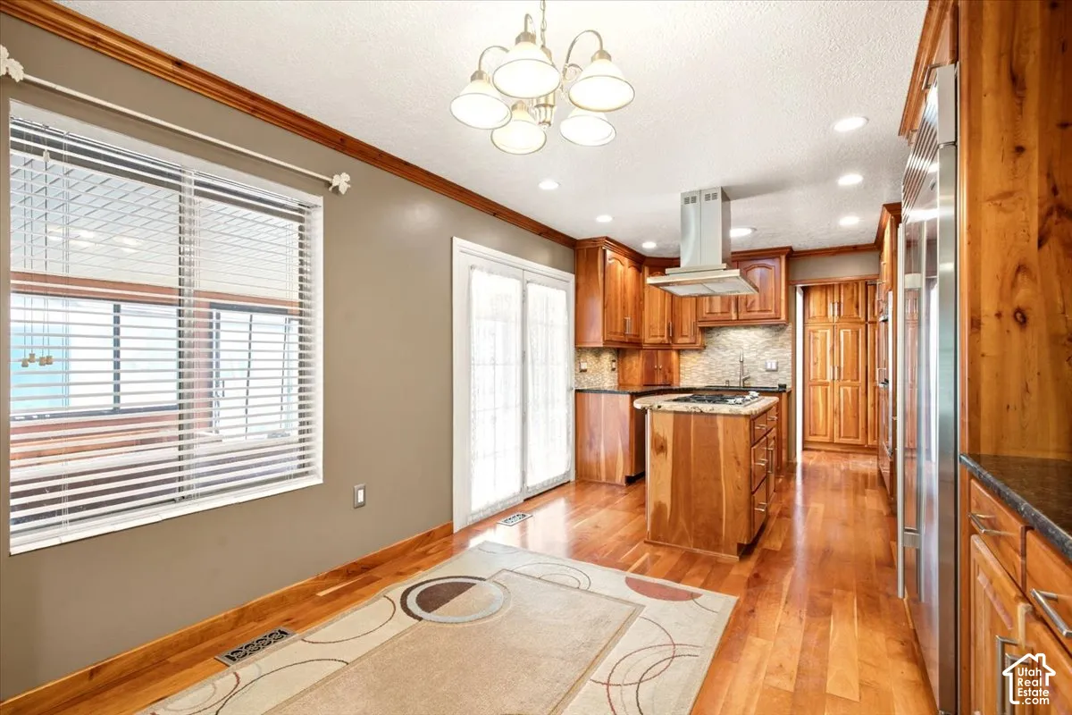 Kitchen featuring custom wood cabinetry, central island with built-in gas cooktop, stainless steel range hood, stone tile backsplash, and wood-finish flooring