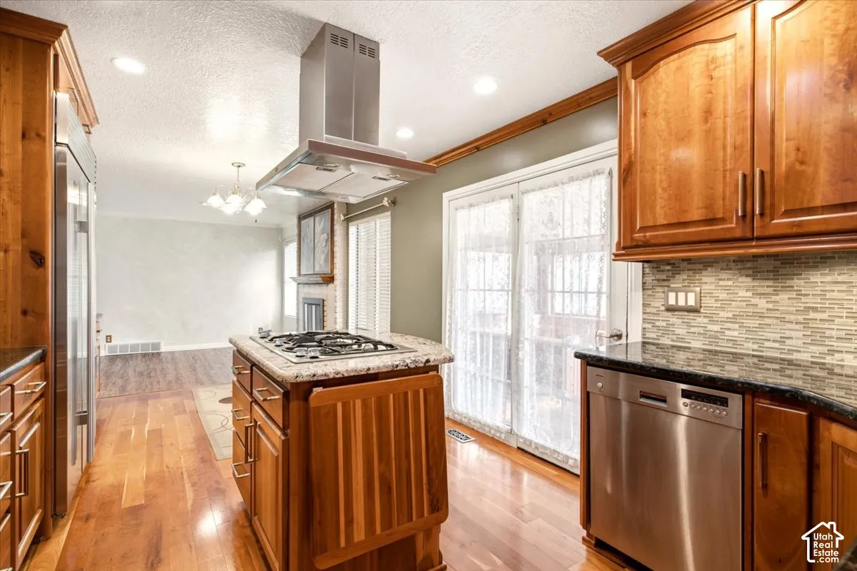 Kitchen featuring custom wood cabinetry, stainless steel appliances, stone countertops, central island with a built-in gas cooktop, and a stainless steel vent hood