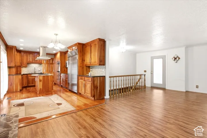 Kitchen featuring custom wood cabinetry, stainless steel professional-grade refrigerator, integrated island with cooktop, light-colored stone backsplash, and wood-finish flooring