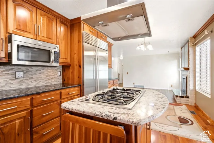 Kitchen featuring natural-finish wood cabinetry, stone countertops, stainless steel appliances, a center island with a built-in gas cooktop, and an overhead stainless steel range hood