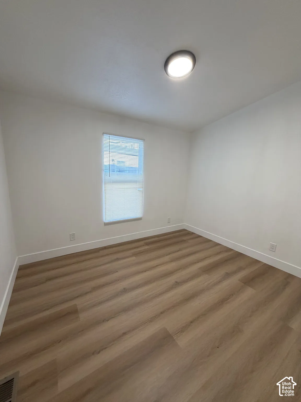 Neutral-toned interior space featuring wood-finish flooring, a single window with blinds, and a flush-mount ceiling light