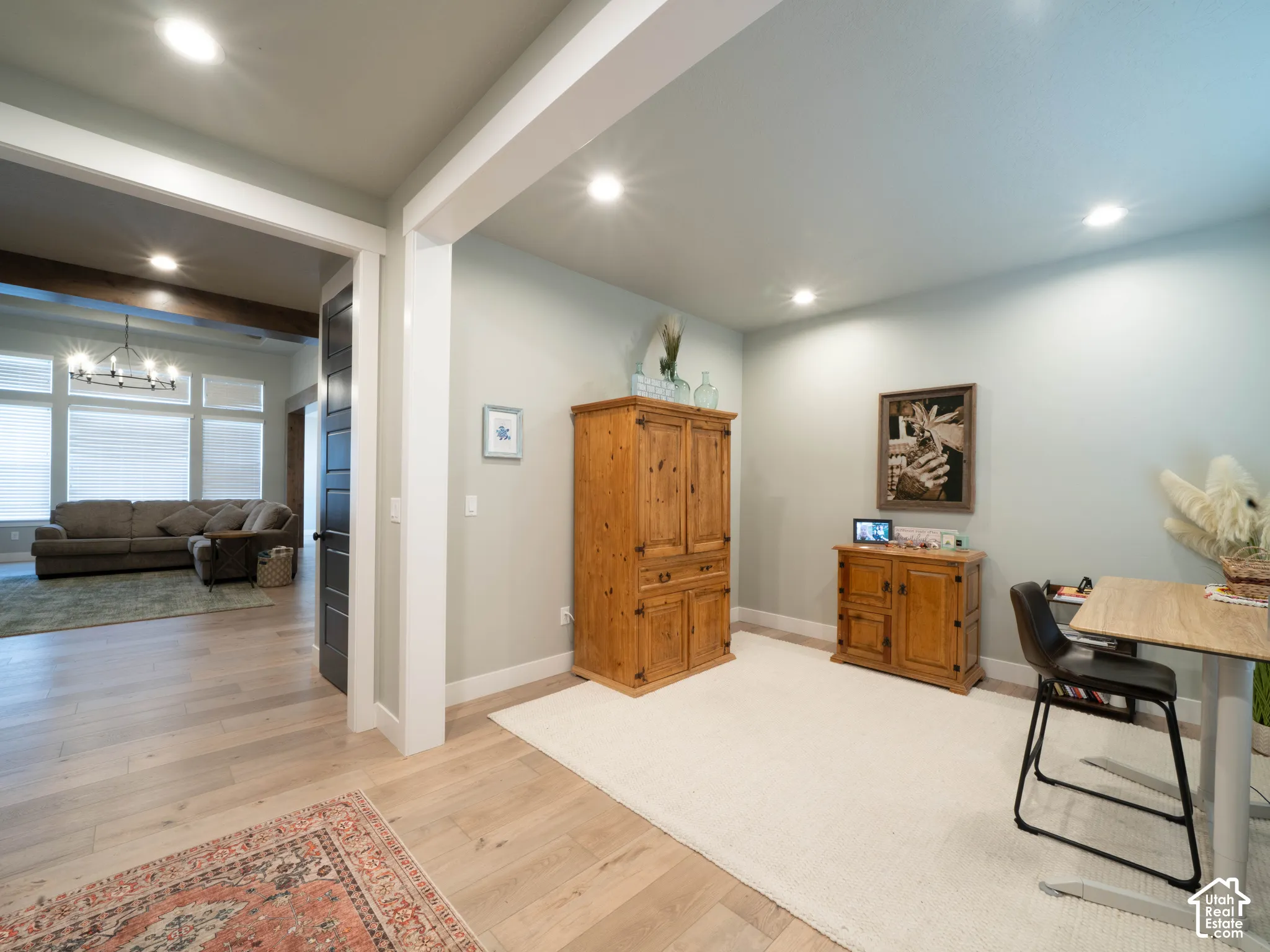 Entry way looking into the front room/office and open living area. Recessed lighting, light wood-finish flooring, open doorway with white trim, dark paneled door, and exposed wood beam.