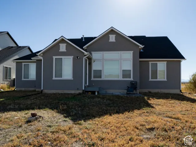 Rear exterior featuring a gray stucco finish, multiple windows, a dark shingle roof, and a concrete patio