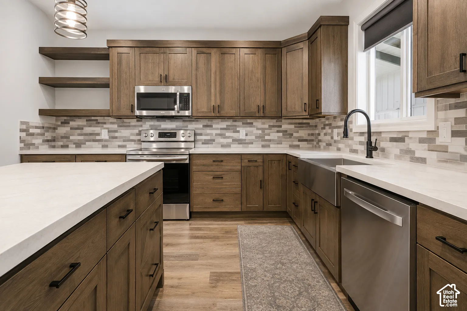 Kitchen featuring wood-finish cabinetry, solid surface countertops, a stainless steel farmhouse sink, open shelving, and a multi-tone tile backsplash