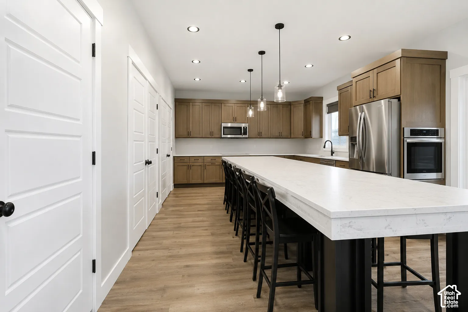 Kitchen featuring wood-finish cabinetry, an expansive island with a light-toned solid surface countertop, stainless steel built-in appliances, recessed lighting, and wood-finish flooring