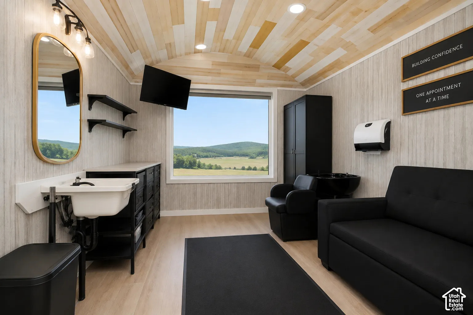 Arched tongue-and-groove wood ceiling, large picture window with rural mountain landscape, vertical-pattern wall paneling, and wall-mounted sink with brass-finish vanity mirror