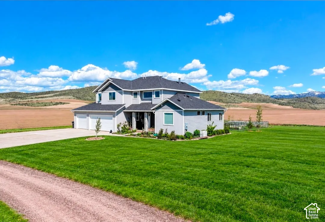 Two-story residence featuring light gray siding, an attached three-car garage, expansive manicured lawn, and mountain backdrop