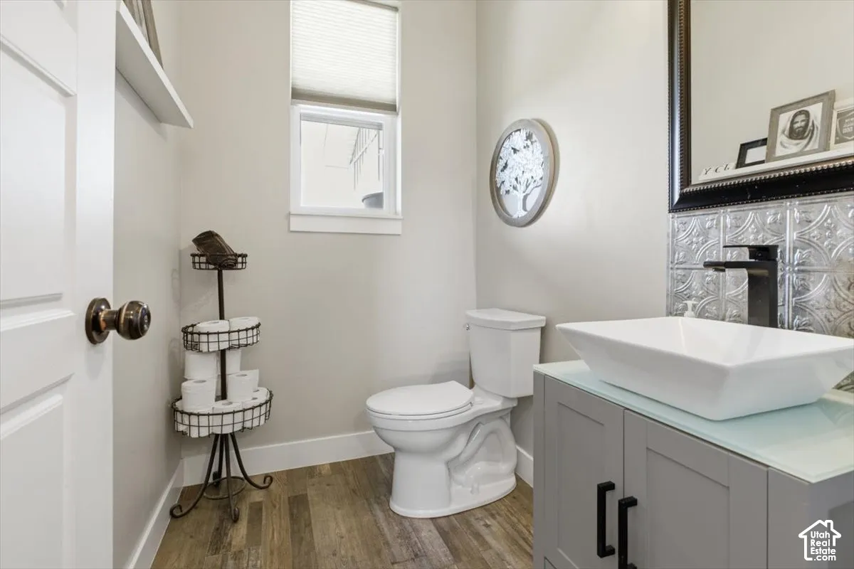 Grey vanity with a glass countertop, rectangular vessel sink, matte black faucet, decorative metal wall tiling, and wood-finish flooring