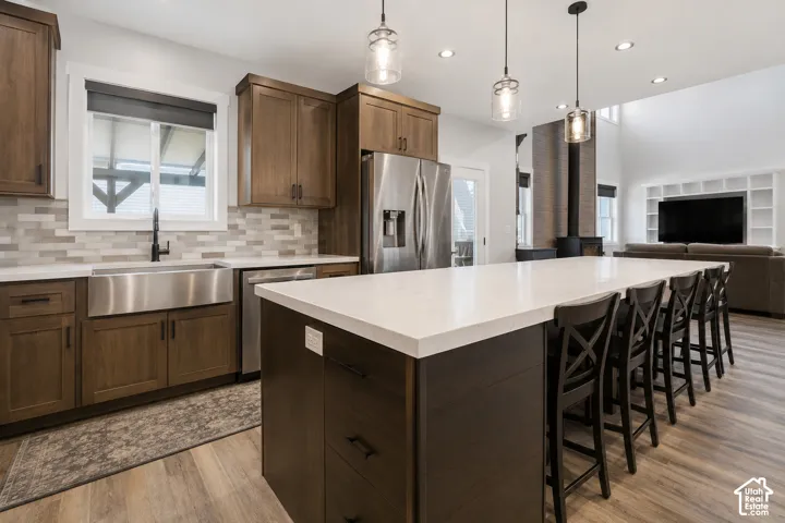 Kitchen featuring medium-tone wood cabinetry, a large central island with a solid-surface countertop, stainless steel apron-front sink, linear tiled backsplash, and pendant lighting