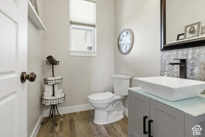 Grey vanity with a glass countertop, rectangular vessel sink, matte black faucet, decorative metal wall tiling, and wood-finish flooring