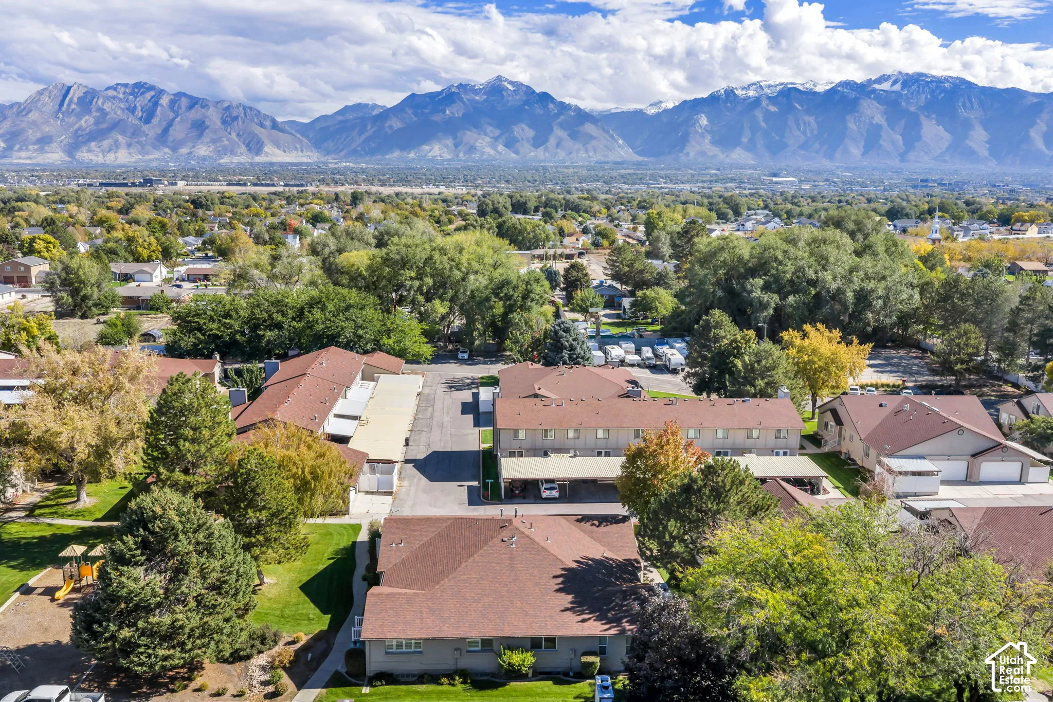 Aerial perspective of the residential complex featuring asphalt-shingled roofing, covered parking structures, mature trees, manicured lawns, and prominent mountain backdrops