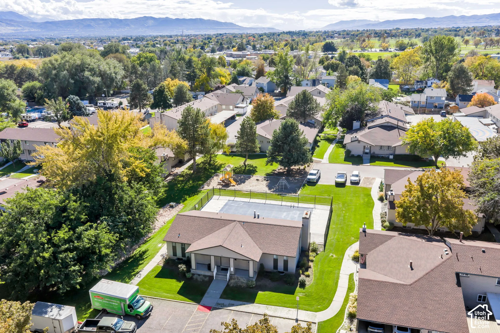Aerial perspective featuring a clubhouse with a fenced outdoor swimming pool, paved parking area, manicured grass landscaping, and mature trees against a mountain backdrop