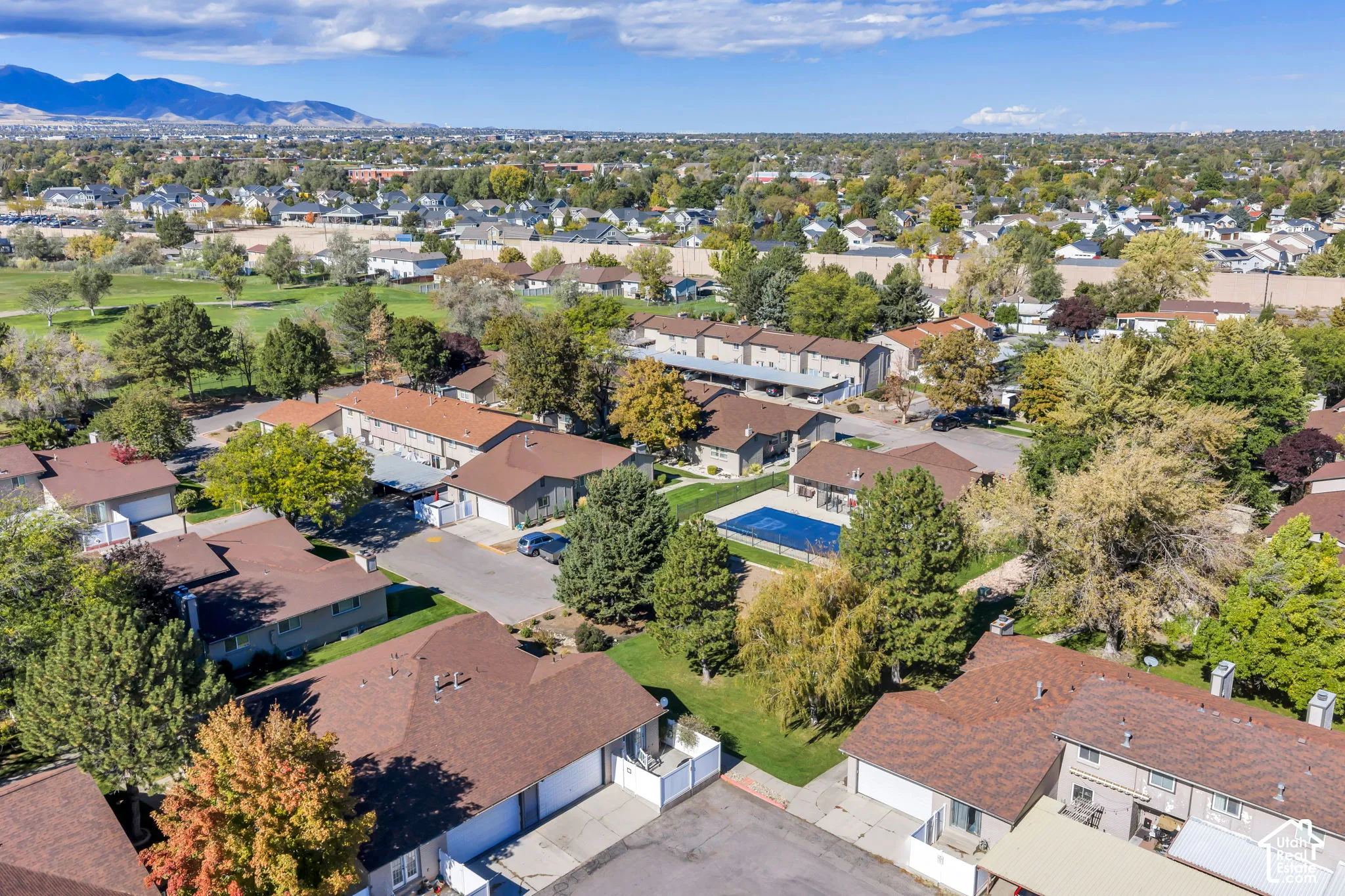 Aerial perspective of a multi-unit residential community featuring brown shingle roofing, integrated garages, a fenced swimming pool, and mature trees with a mountain backdrop