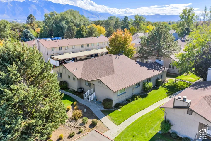 Multi-family structure with a multi-gabled asphalt shingle roof, exterior concrete walkway, covered parking area, and manicured green space set against a mountain backdrop