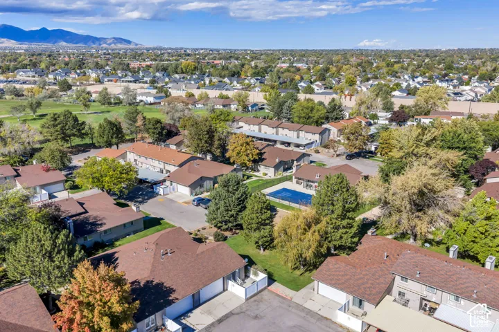 Aerial perspective of a multi-unit residential community featuring brown shingle roofing, integrated garages, a fenced swimming pool, and mature trees with a mountain backdrop