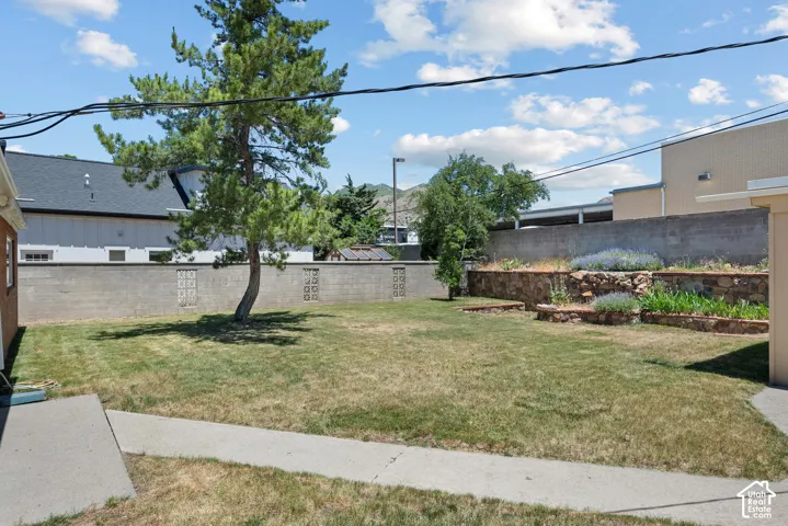 Backyard featuring a concrete walkway, stone-retaining walls, tiered garden beds, decorative masonry block perimeter fencing, and a mature evergreen tree