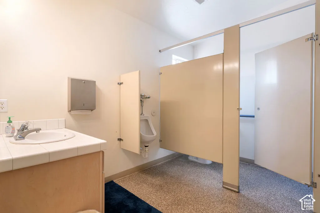 Restroom featuring a vanity with tiled countertop, stainless steel paper towel dispenser, wall-mounted urinal, and metal privacy partitions