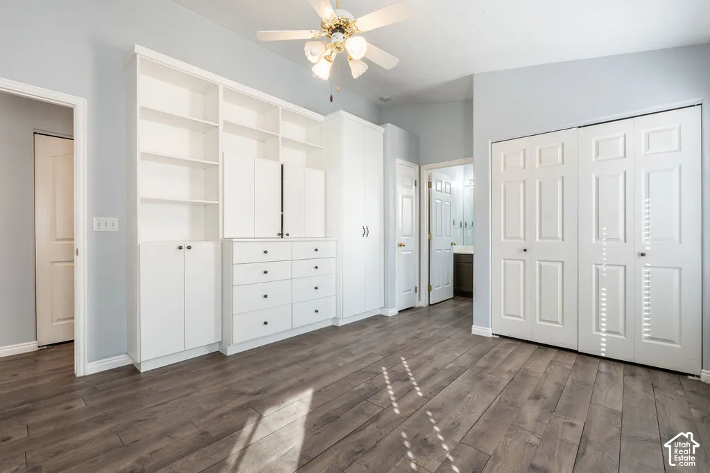Room featuring wood-finish flooring, extensive built-in white cabinetry with shelving and drawer storage, double-door closets with six-panel detailing, and a ceiling-mounted fan with light fixture