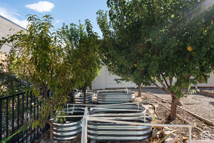 Exterior garden area featuring multiple elevated galvanized steel planting beds, integrated irrigation piping, mature trees, and white vinyl privacy fencing