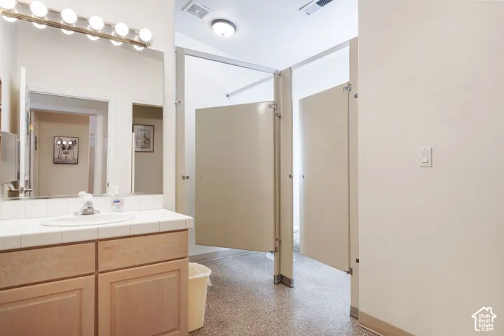 Wood-finish cabinetry, tile-surround vanity countertop with an integrated basin, wall-mounted multi-bulb lighting fixture, and industrial-style partitioned stalls