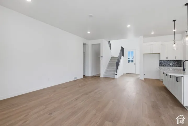 Open-concept interior featuring light wood-finish flooring, white cabinetry with stone countertops, recessed lighting, glass pendant fixtures, and a staircase with a black metal railing