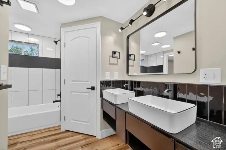 Dual vessel sinks atop a floating vanity with dark cabinetry, complemented by black subway tile backsplash, a rectangular framed mirror with integrated lighting, and wood-finish flooring