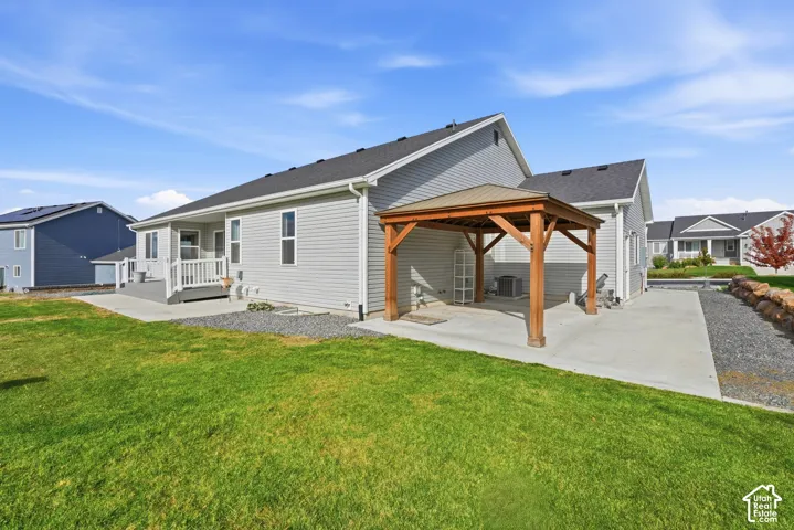 Rear exterior featuring vinyl siding, a covered wooden pavilion, an expansive concrete patio, a raised deck with white railing, and a boulder-lined retaining wall