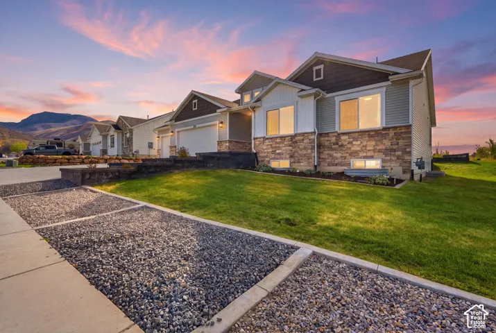 Multi-level residential exterior featuring stone veneer siding, horizontal lap siding, a concrete driveway, and a gravel parking area against a mountain backdrop