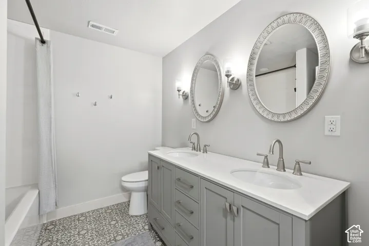 Dual-sink vanity with quartz countertop, grey cabinetry, patterned tile flooring, and oval wall mirrors