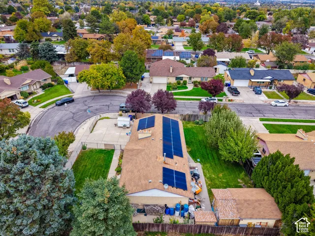 Aerial perspective of a single-story residence featuring a shingle roof equipped with a solar panel array, a concrete driveway, a chain-link fenced perimeter, and established landscaping