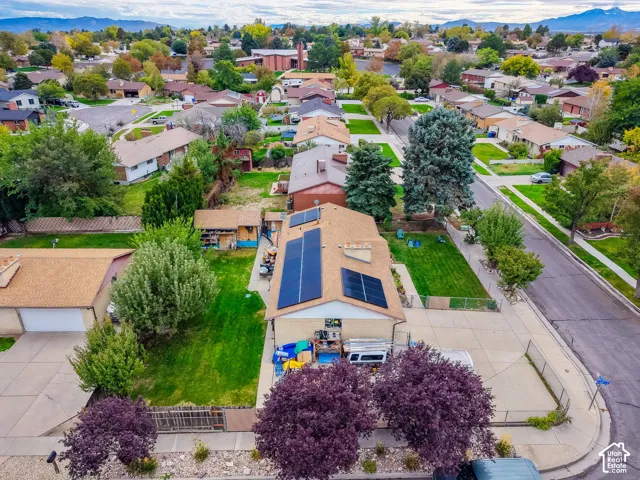 Aerial perspective of a single-story residence featuring a shingle roof equipped with solar panel arrays, a concrete driveway, a chain-link fenced yard, and mountain scenery in the distance