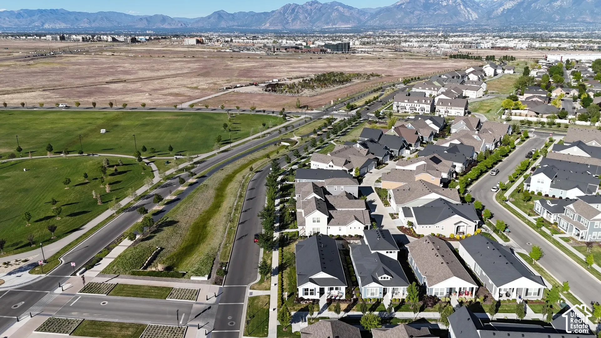 Aerial perspective of a residential community featuring multi-level single-family homes, paved streets with sidewalks, and an expansive open-space park area, framed by a prominent mountain range backdrop