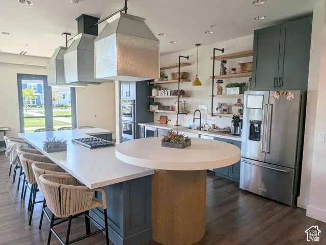 Kitchen featuring a dual-level island with a solid surface countertop, custom dark-toned cabinetry, stainless steel appliances, metal pendant lighting, and open wood shelving