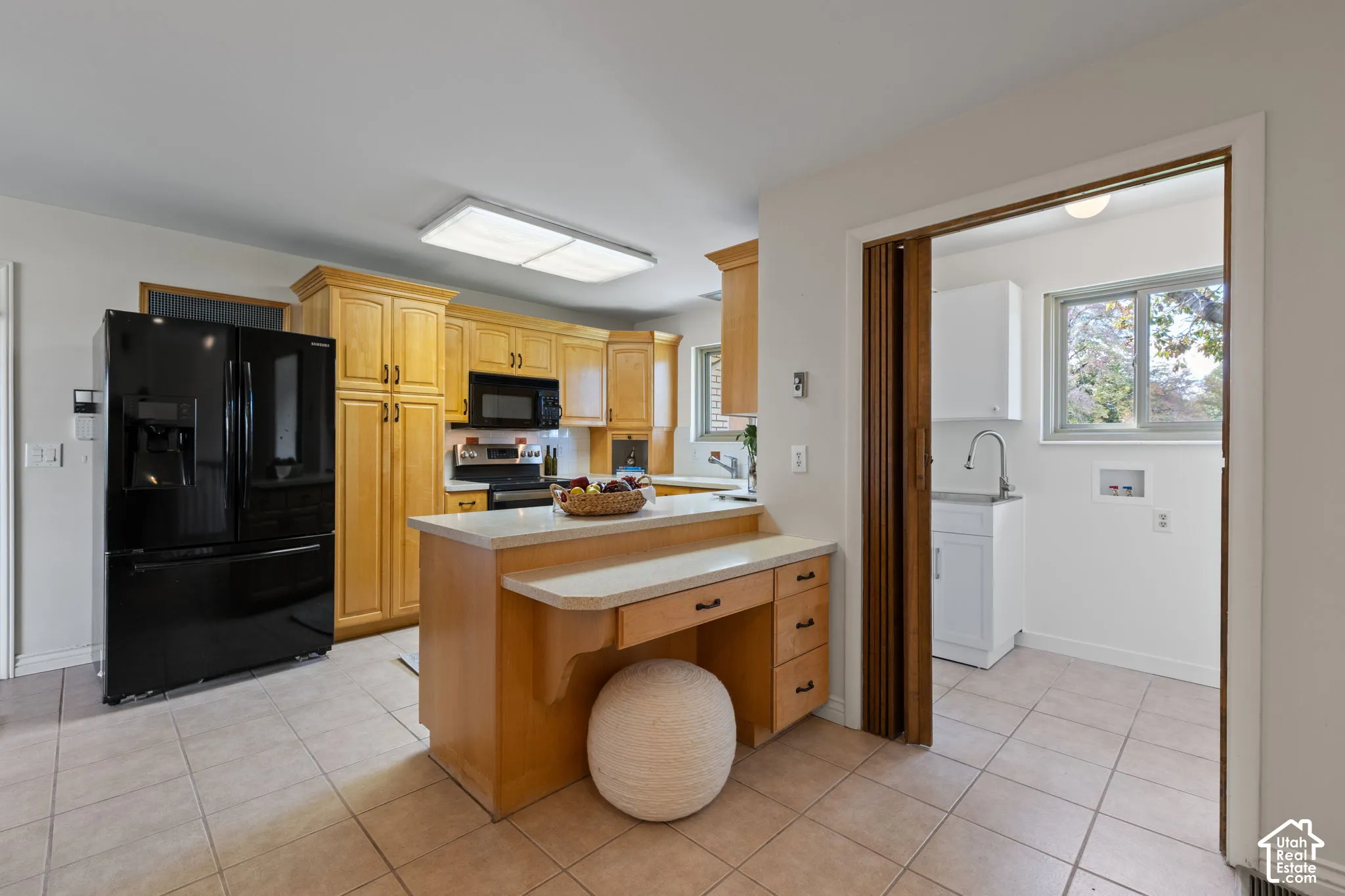 Kitchen featuring honey-tone wood cabinetry, central island with integrated desk, black refrigerator, neutral tile flooring, and bi-fold door access to a secondary workspace with white cabinetry and utility sink