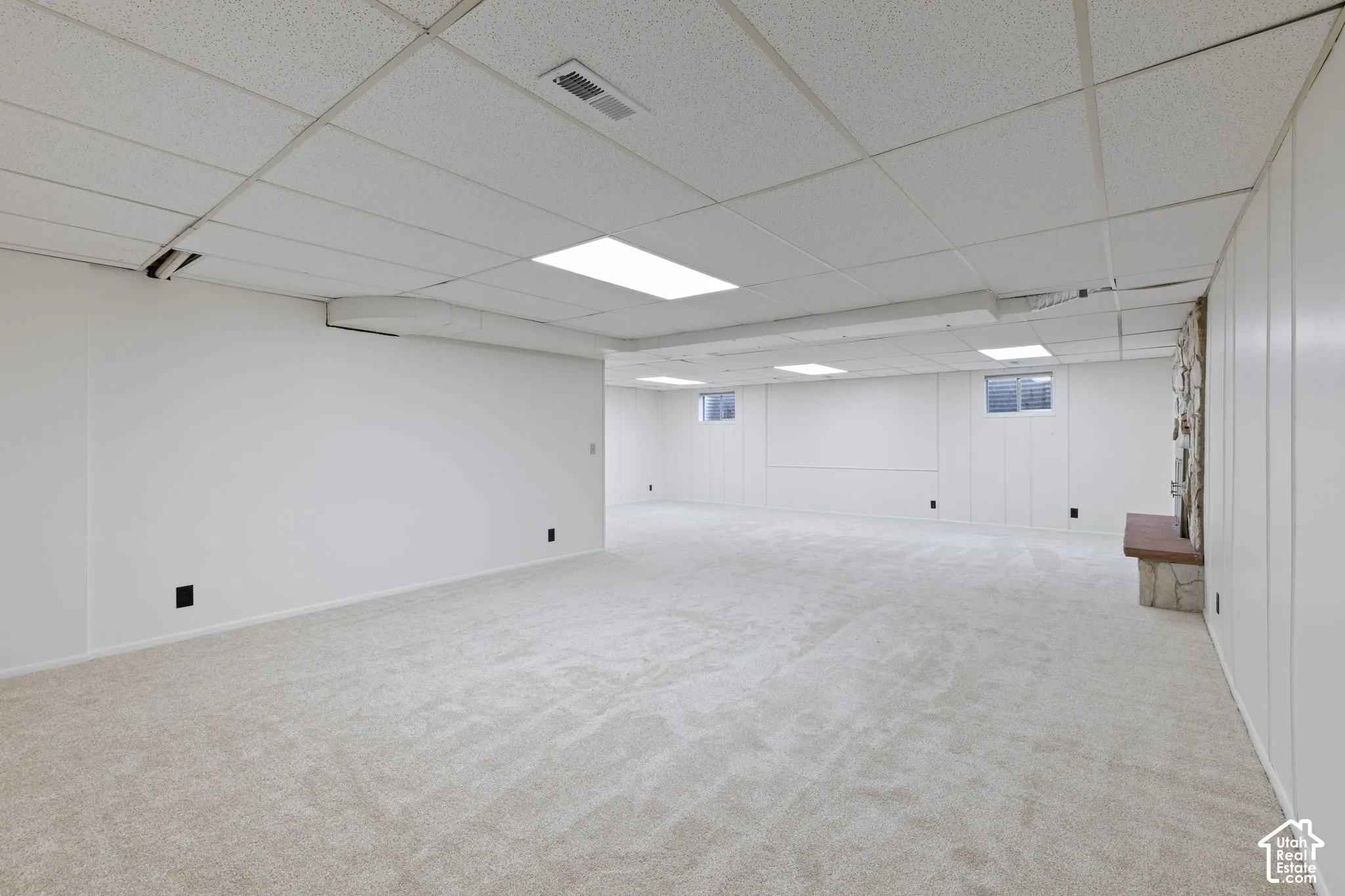 Expansive open-concept space featuring wall-to-wall carpeting, a drop ceiling with integrated lighting, paneled walls, and a stone-accented hearth