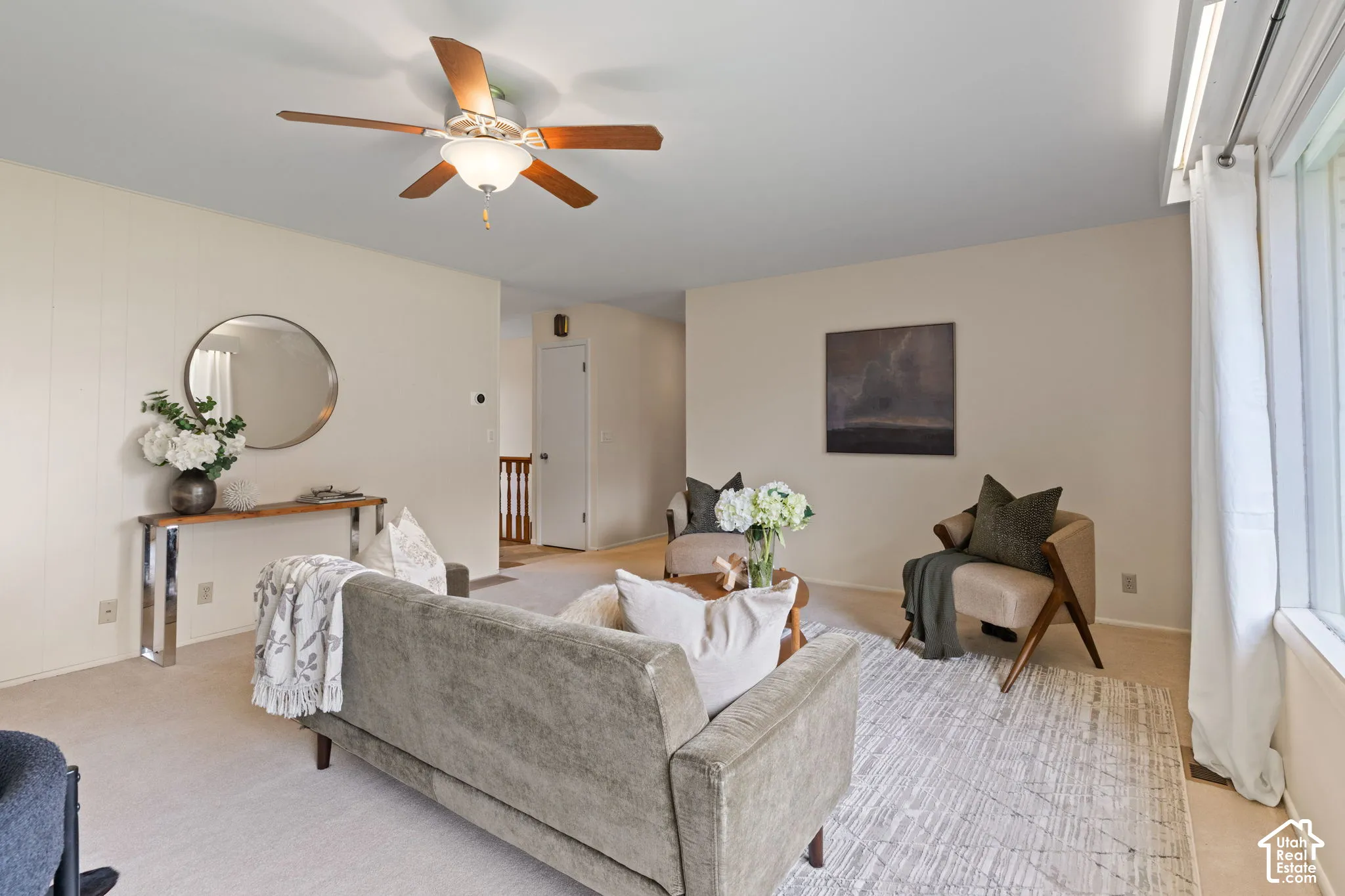 Neutral-toned room featuring a central ceiling fan with integrated lighting, expansive window with hardware, and carpeted flooring