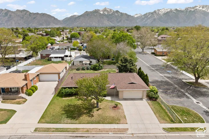 Aerial perspective of a single-level residence featuring a concrete driveway, two-car garage, asphalt shingle roof, and mature landscaping, framed by a mountain backdrop