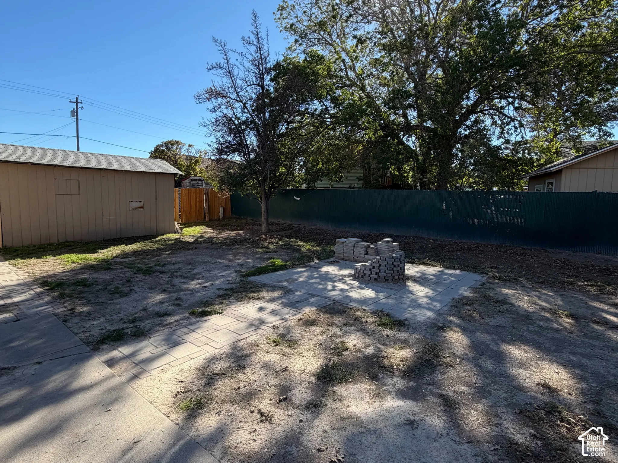 Backyard space featuring a detached storage shed, paver stone patio, and mature trees enclosed by metal fencing