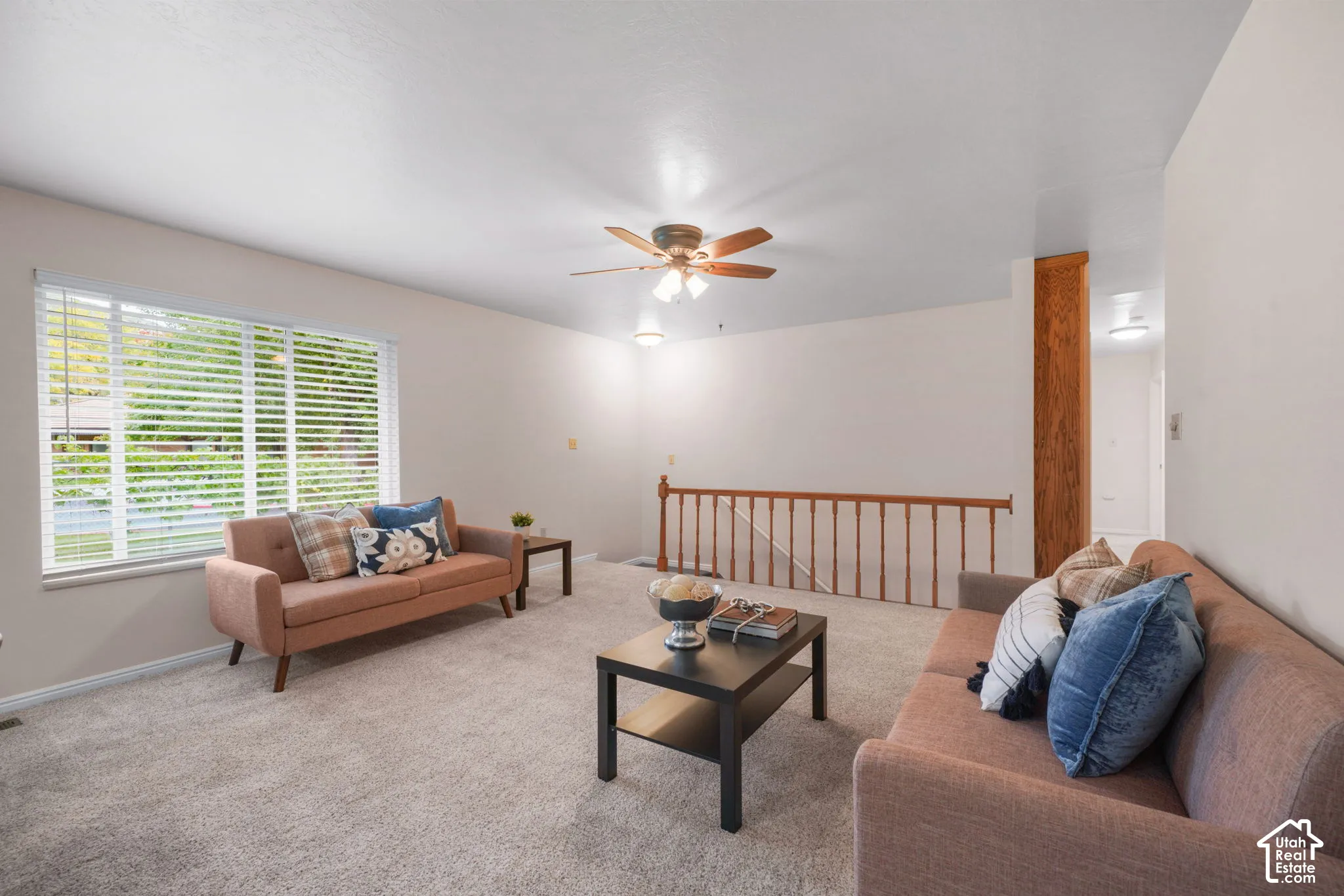 Neutral-toned carpet, large window with horizontal blinds, ceiling fan with integrated lighting, and wooden stair railing to basement