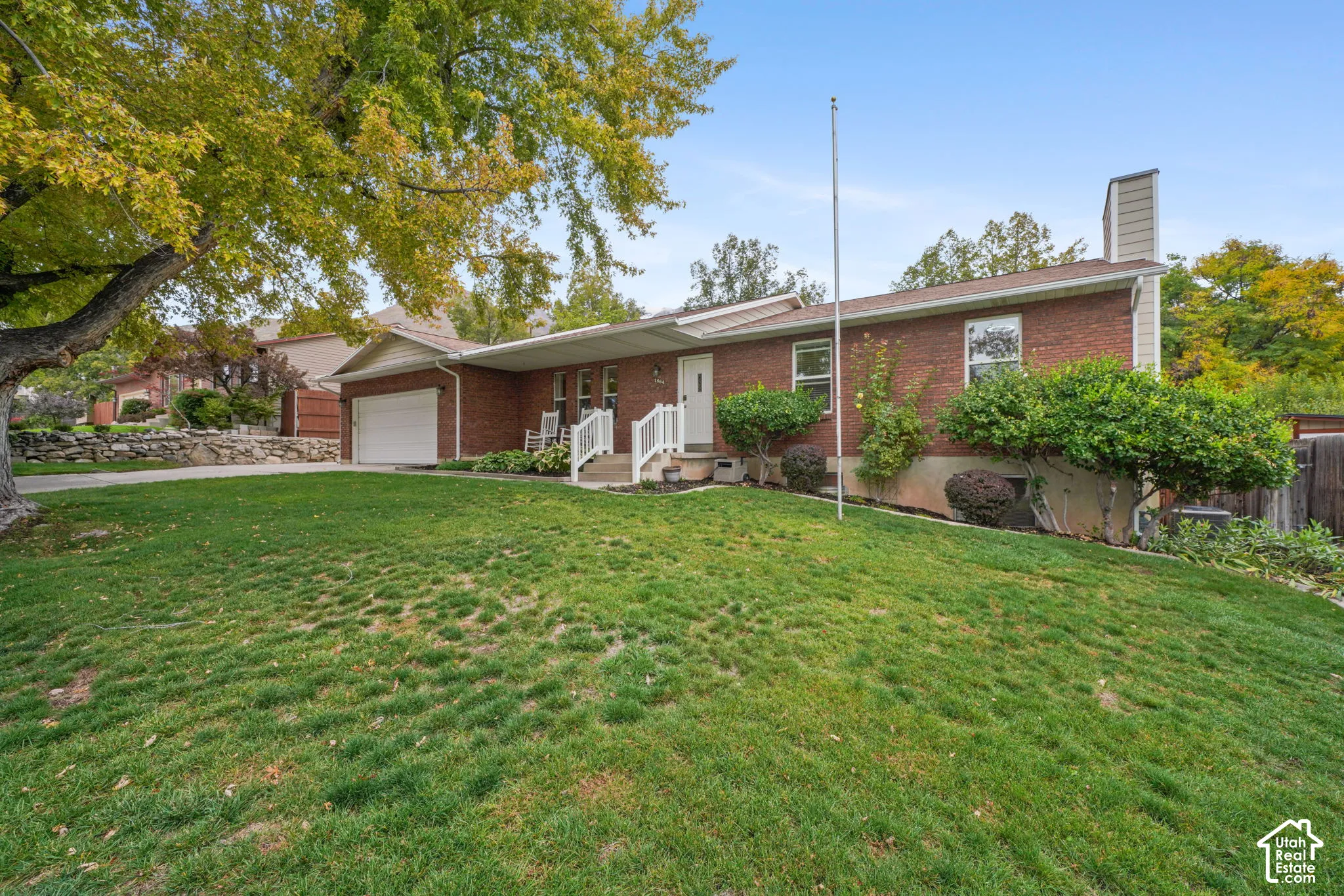 Ranch-style exterior featuring red brick masonry, an attached two-car garage, mature landscaping, a stone retaining wall, and white exterior railings
