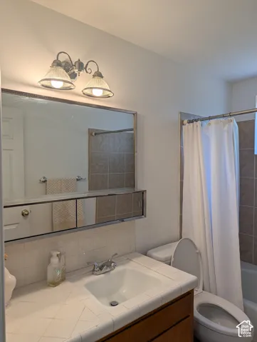 Bathroom featuring a tile-countertop vanity, dual-door medicine cabinet, chrome dual-handle faucet, two-light fixture with ribbed glass shades, and a tiled shower surround