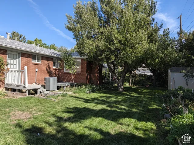 Backyard area featuring red brick exterior, elevated wood deck with white balusters, expansive lawn, mature tree canopy, and detached storage shed