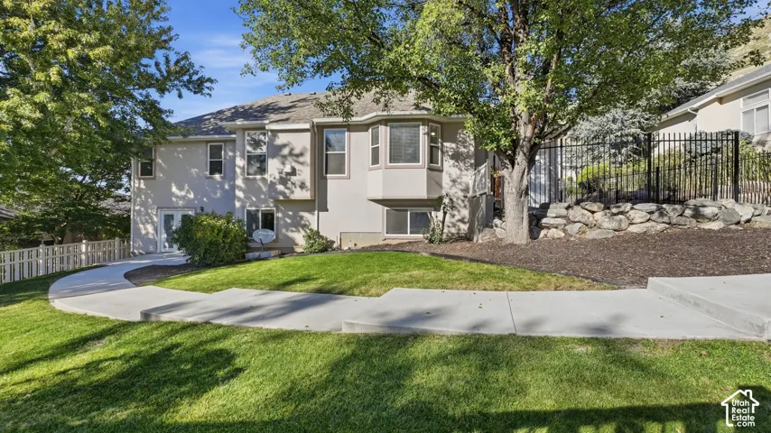 Multi-level stucco exterior with a bay window, concrete walkways, mature trees, and tiered landscaping with decorative rock retaining walls
